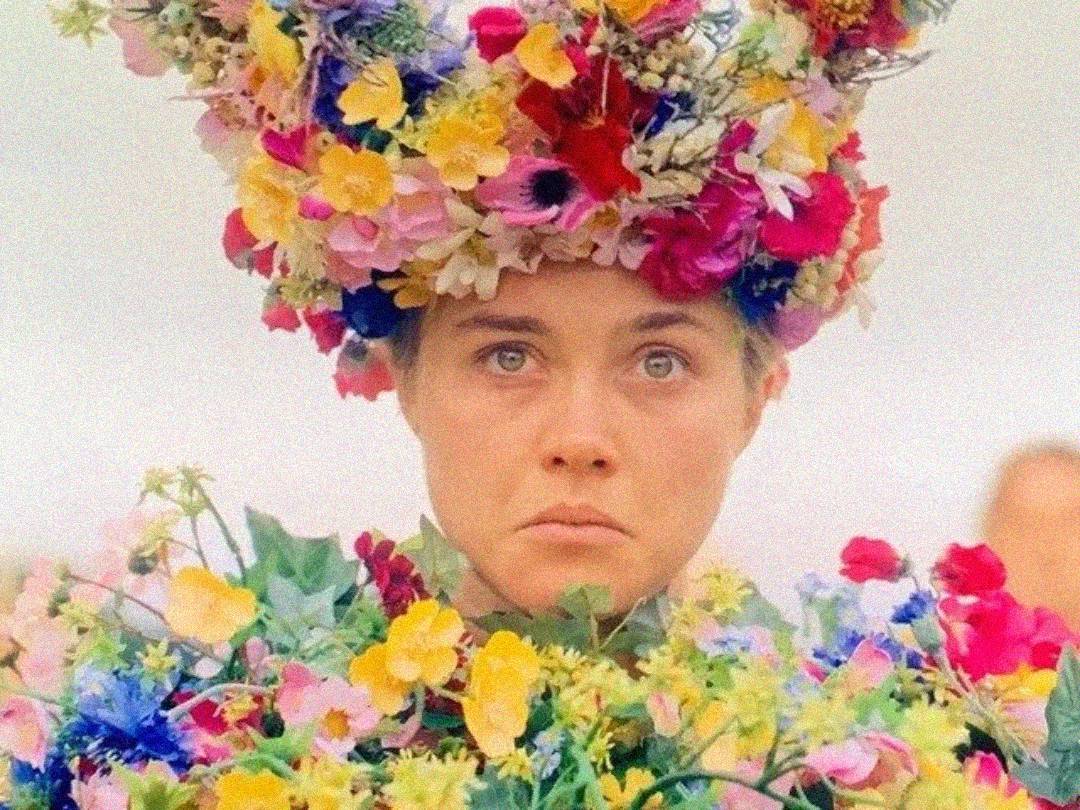 A person with a serious expression wears an elaborate crown and dress made entirely of colorful flowers, set against a pale, blurred background.