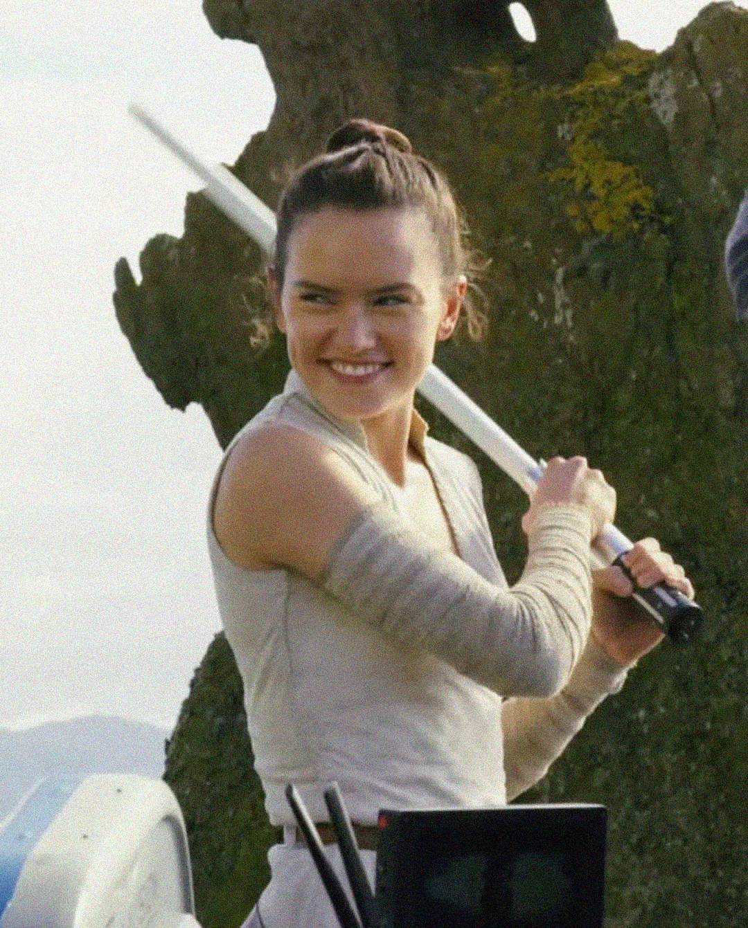 A woman in beige clothing holds a lightsaber with both hands while smiling, standing outdoors near a large rock formation.