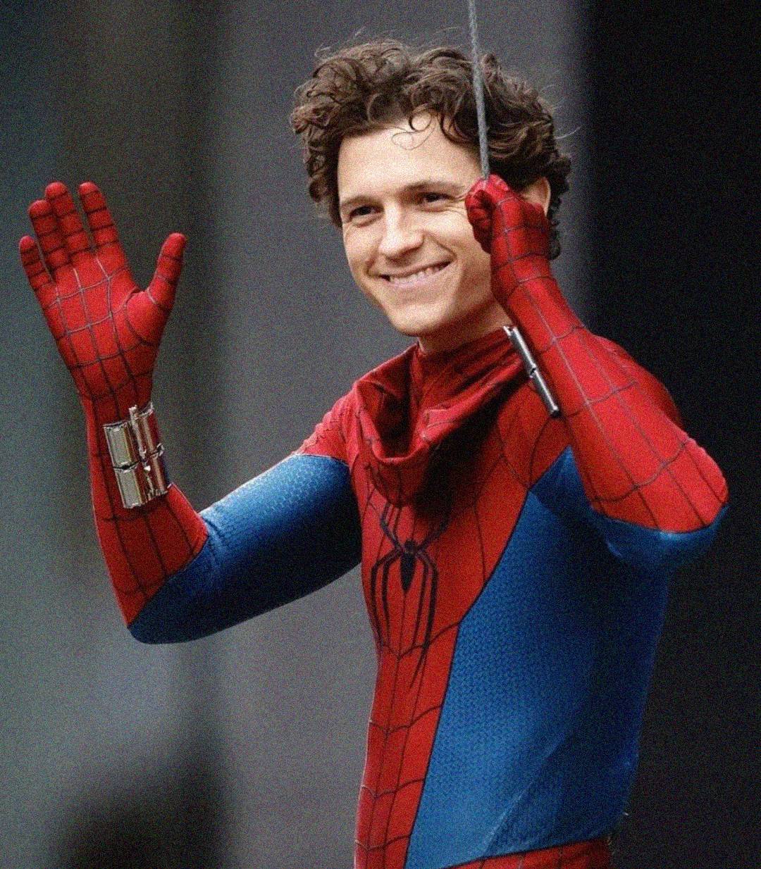 A person in a Spider-Man costume smiles and waves while holding onto a web-like rope with one hand. The suit is red and blue with black web patterns. The background is blurred.