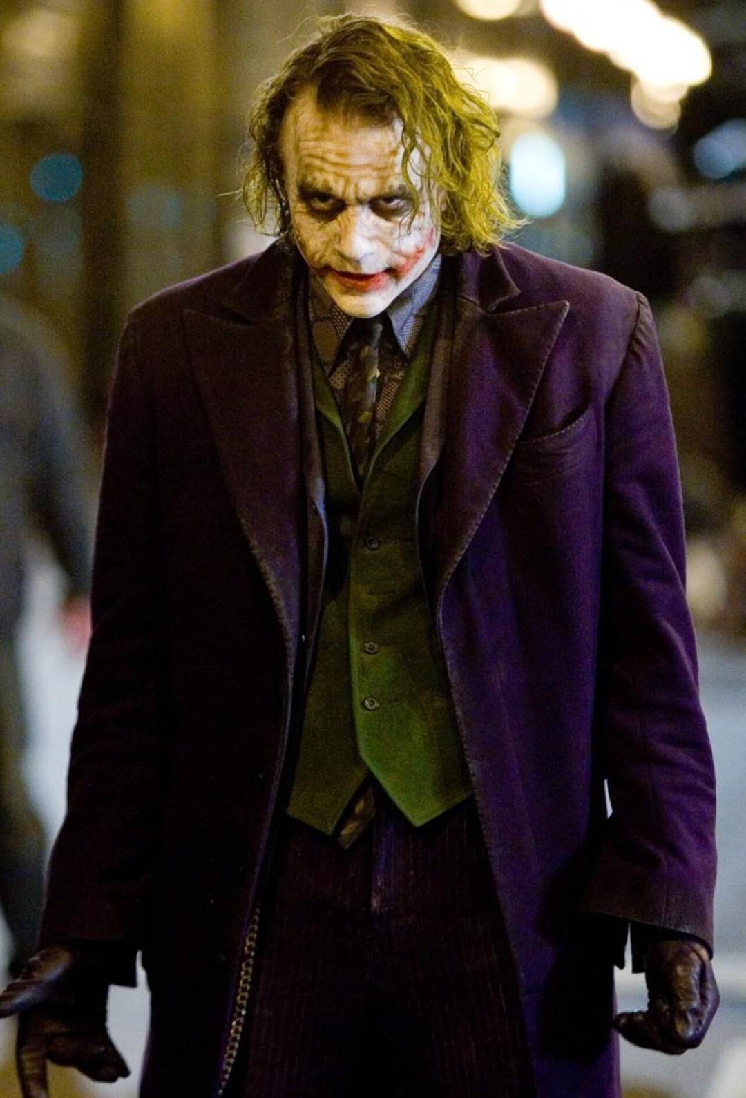 A person dressed as the Joker from "The Dark Knight" wears smeared white face paint, dark eye makeup, red lipstick, green hair, a purple coat, green vest, and dark gloves, standing with a menacing expression.