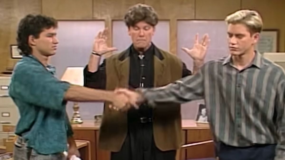 Three men are indoors; two are shaking hands in the foreground, while the third stands behind them with both hands raised, appearing to react dramatically to the handshake. Office furniture is visible in the background.