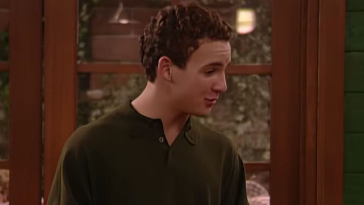 A young man with short curly hair wearing a dark green shirt stands indoors, looking to his right. Wooden framed windows and blurred lights are visible in the background.
