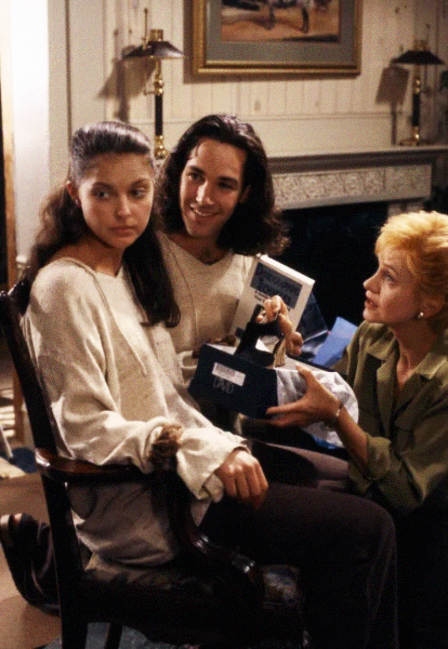 Three people gather indoors; one woman sits in a chair looking away, another sits beside her smiling and holding a box, while a third, kneeling, offers a videocassette tape, all appearing engaged in conversation.