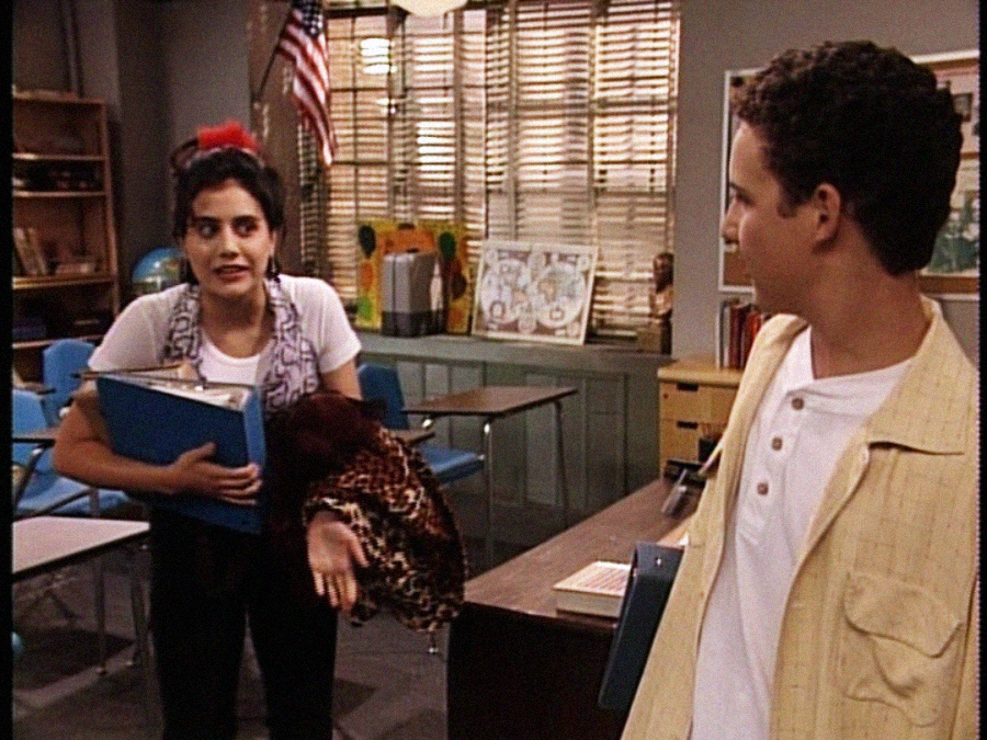A young woman holding books and a jacket gestures while talking to a young man in a classroom with desks, a U.S. flag, and blinds covering the windows in the background.
