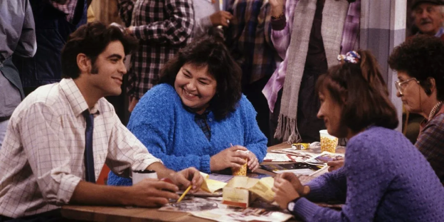 A group of people sit around a table, smiling and talking. One man in a white shirt is smiling at a woman in a blue sweater, who sits beside two other women. The table is covered with papers and drinks; people stand in the background.