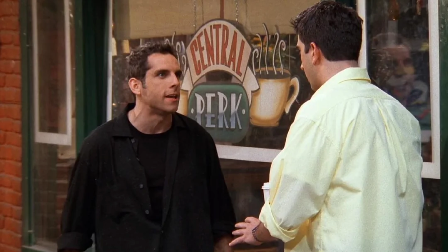 Two men are having an intense conversation outside Central Perk café; one looks upset, wearing black, while the other, holding a coffee cup, faces him, wearing a light yellow shirt.