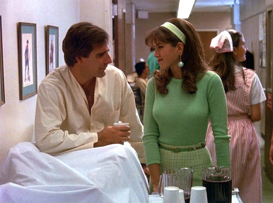 A man in a cream shirt and a woman in a green top stand talking in a hallway, with a hospital setting suggested by uniforms and medical equipment. Two other women in nurse uniforms are seen in the background.