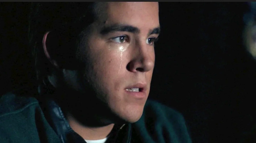 A young man with short brown hair stares ahead with a somber expression as a single tear runs down his cheek, illuminated by soft lighting against a dark background.