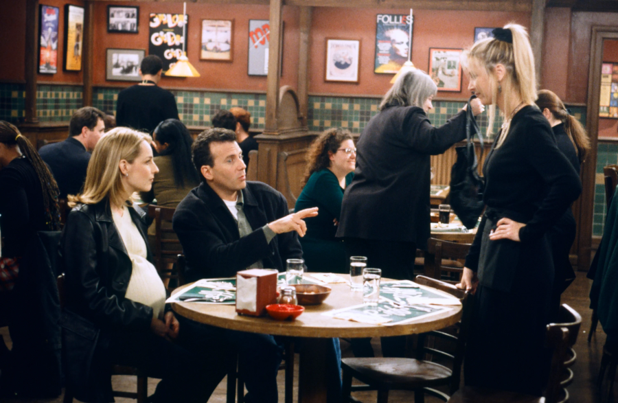 A pregnant woman and a man sit at a round cafe table, talking to a blonde waitress standing beside them. The cafe is busy, with other patrons seated and posters on the wall in the background.