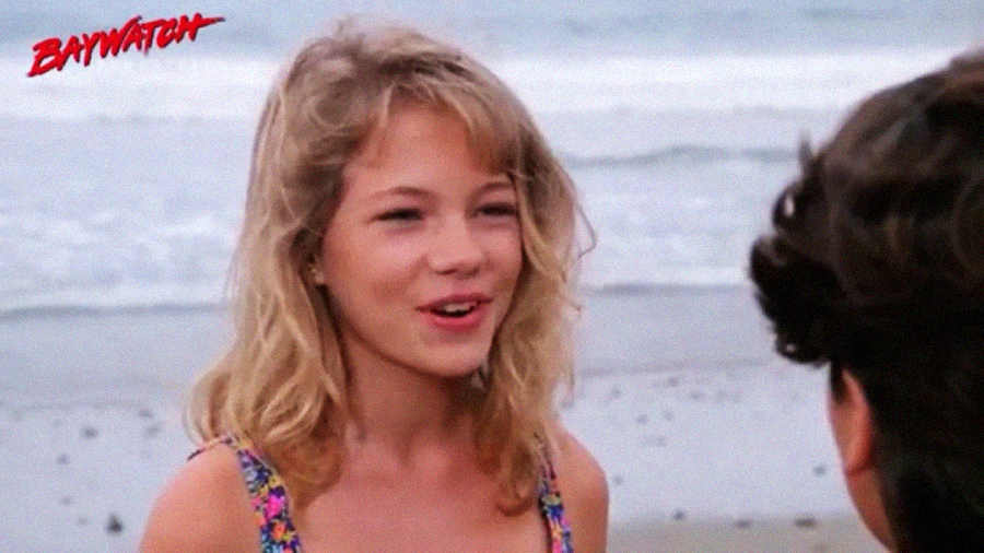 A young woman with wavy blonde hair stands on a beach, smiling and talking to someone whose back is to the camera. The ocean is in the background, and the "Baywatch" logo is in the top left corner.