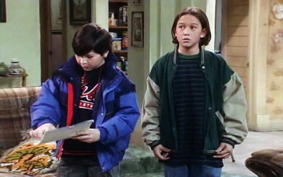 Two boys stand in a living room; one with dark hair and a blue jacket looks down at papers, while the other with longer hair and a green jacket looks ahead with a concerned expression.