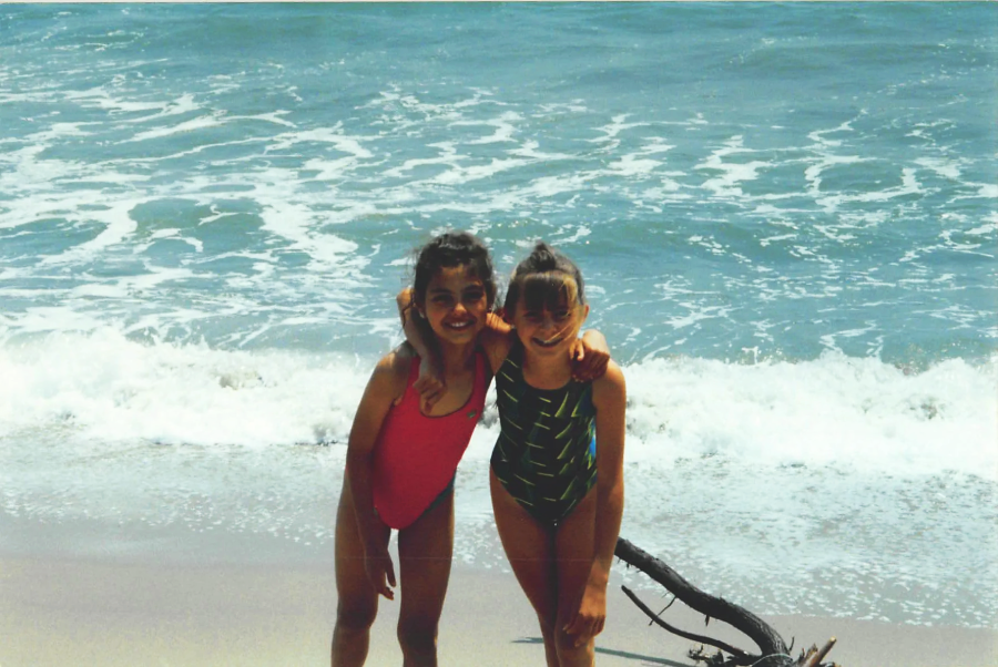 Two young girls in swimsuits stand on a sandy beach with waves crashing behind them. One girl wears a red swimsuit, the other a green striped one. They are smiling and have their arms around each other. A piece of driftwood lies nearby.