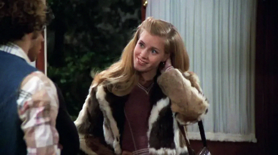 A young woman with long blond hair, wearing a fur coat, smiles and tucks her hair behind her ear while talking to someone indoors near a window with white curtains.