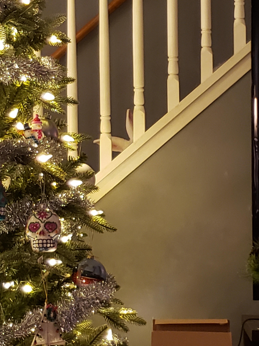 A white and brown cat peeks through the railing of a staircase behind a lit Christmas tree decorated with tinsel and ornaments.