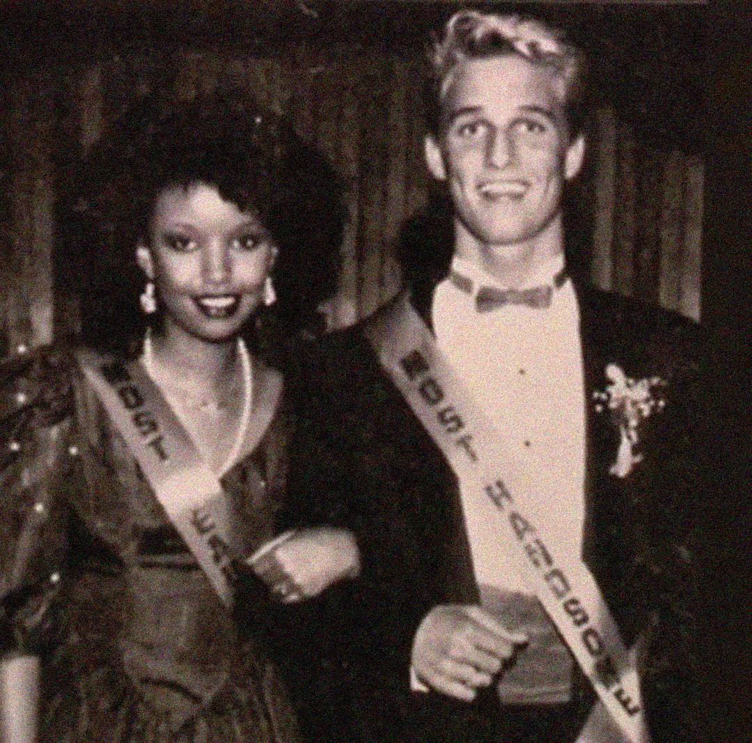 A young woman and young man wearing formal attire and sashes pose together at an event. The woman has curly hair and wears a dress and earrings; the man is in a tuxedo. Both are smiling. The photo is black and white.