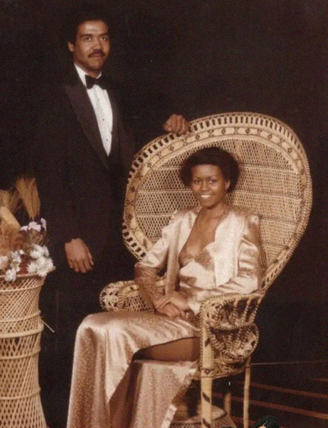 A man in a tuxedo stands beside a woman seated in a peacock chair, wearing a gold evening gown with a deep neckline. They both pose formally against a dark backdrop with a wicker table and a floral arrangement.