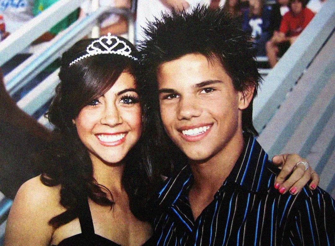 A teenage girl in a black dress and tiara smiles next to a teenage boy in a striped shirt. They are posing together with their arms around each other in a crowded bleacher setting.