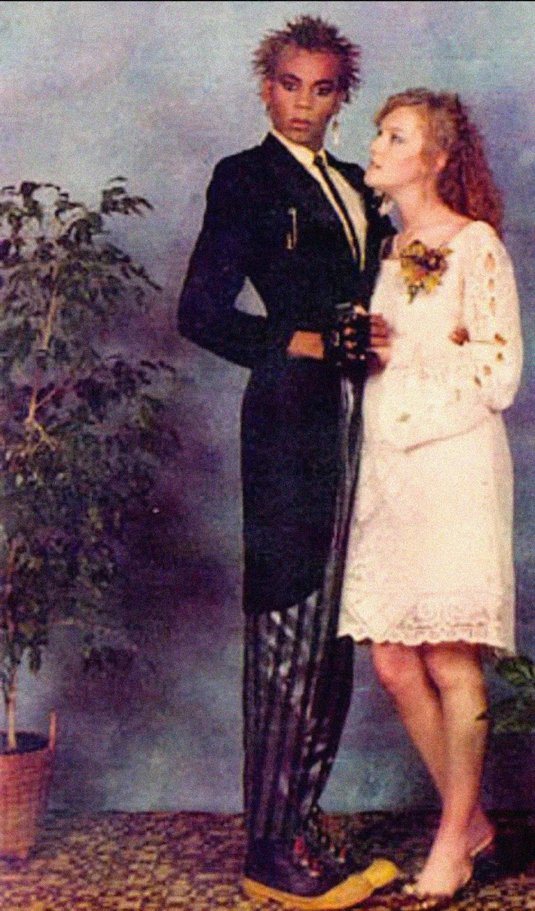 Two people in contrasting prom outfits pose together. One wears a black tailcoat, striped pants, spiked hair, and boots; the other wears a white lace dress with a corsage and curly hair. They stand beside a potted plant against a studio backdrop.