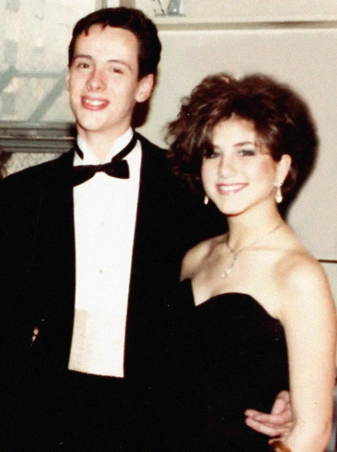 A young man in a tuxedo stands with a young woman in a black strapless dress; both are smiling and posing for a formal photo, likely at a prom or formal event.