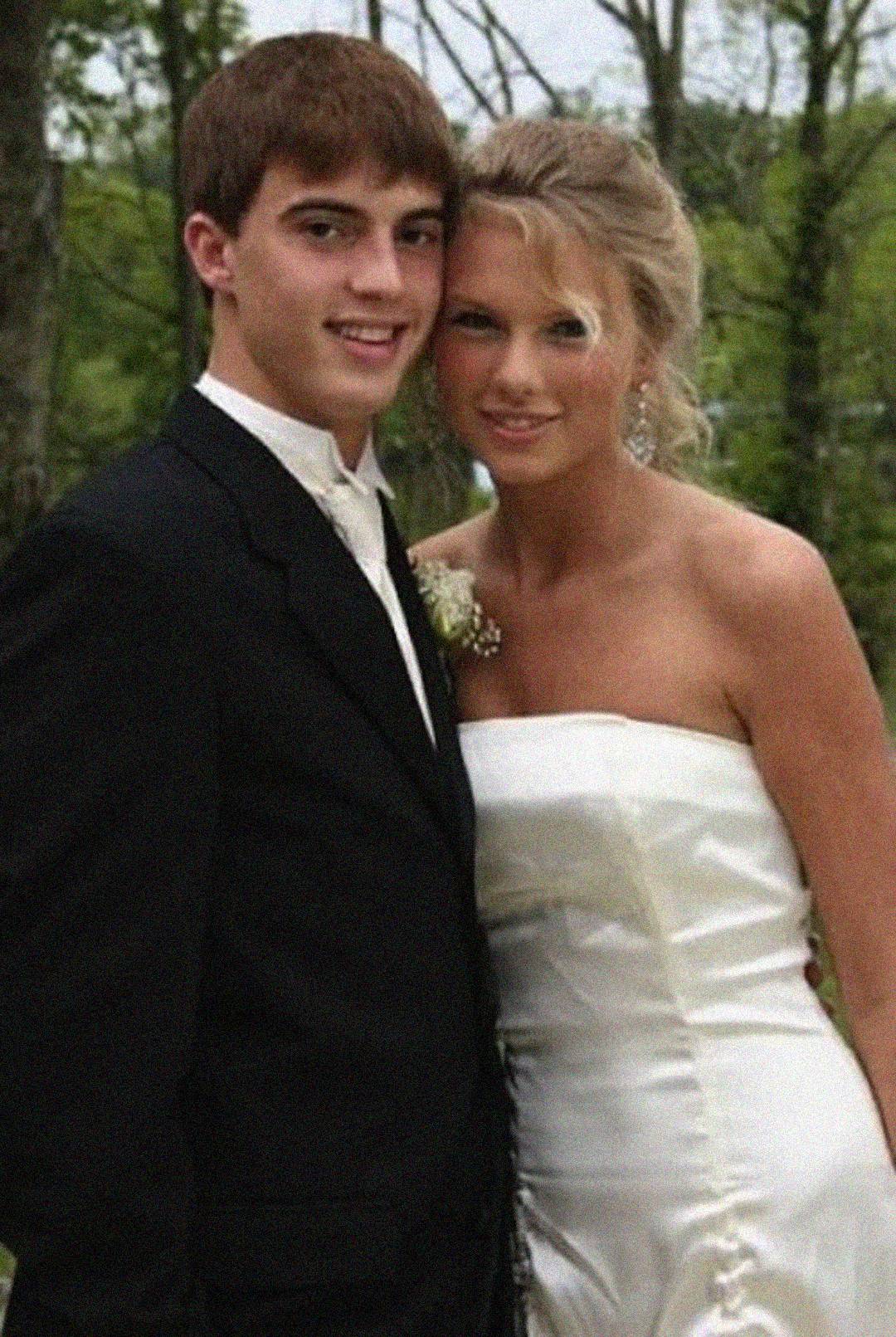 A young man in a black suit poses next to a young woman in a strapless white dress, both smiling outdoors with green trees in the background.