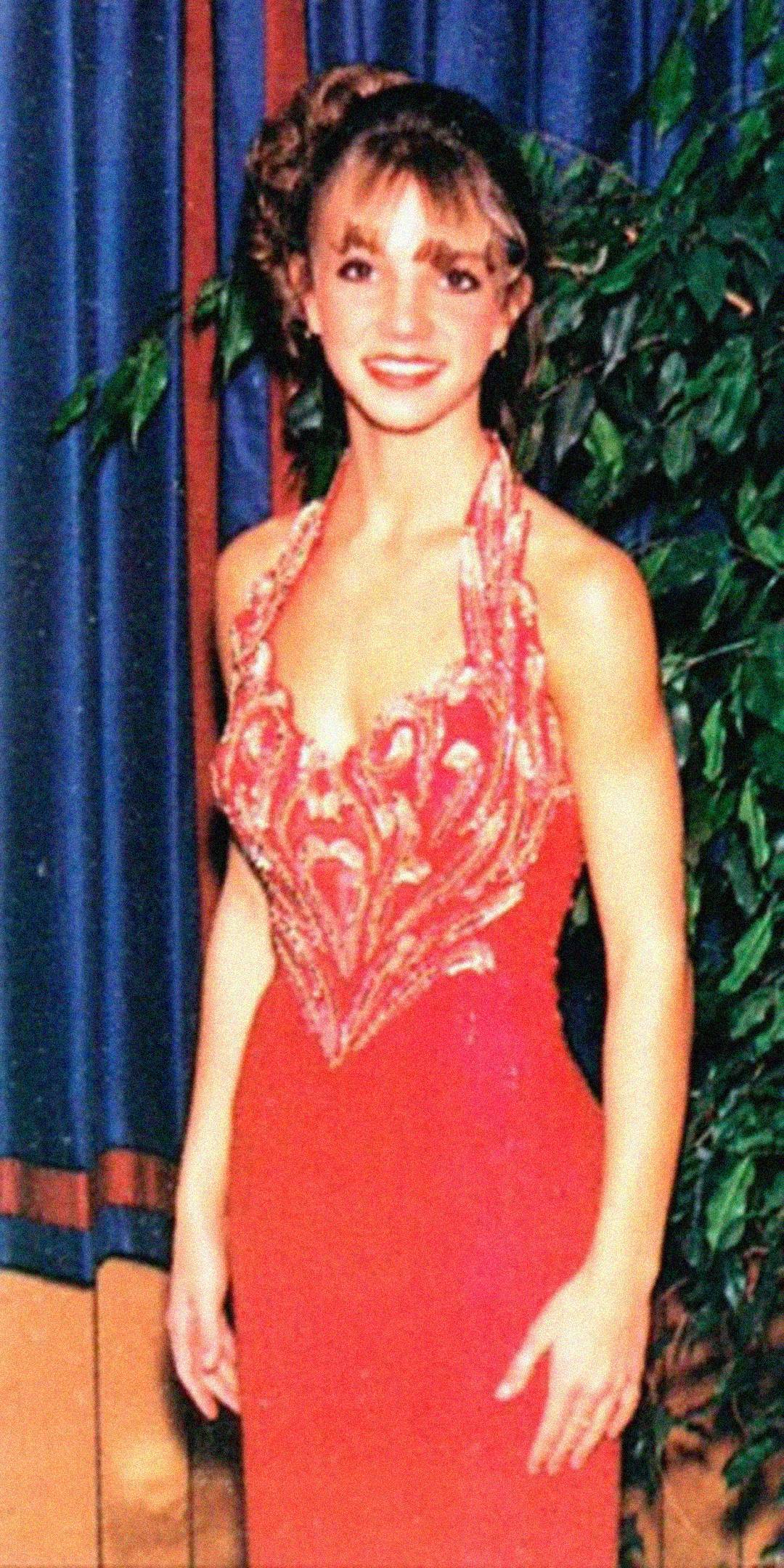 A young woman in a red, embellished halter dress stands indoors, smiling at the camera, with a blue curtain and leafy green plant in the background.