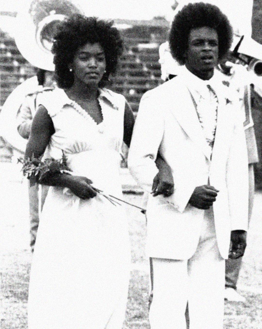 A young woman in a formal dress holds a flower and walks arm-in-arm with a young man in a light-colored suit at an outdoor event, with a marching band playing instruments in the background. The image is black and white.