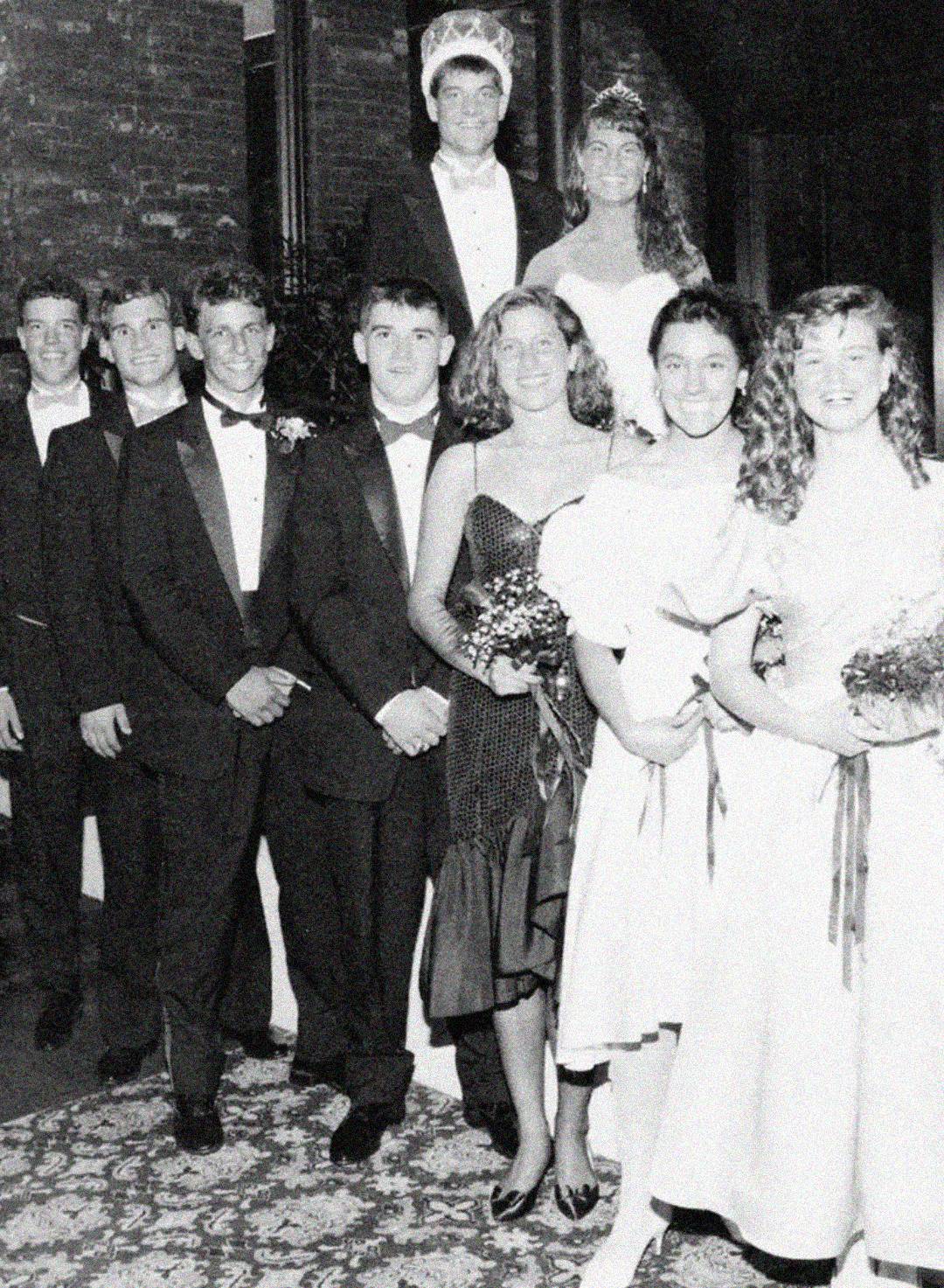 A black-and-white photo of a group of young people in formal attire, with some wearing tuxedos and others in dresses. Two are standing elevated above the group, one wearing a crown, possibly at a prom or homecoming event.