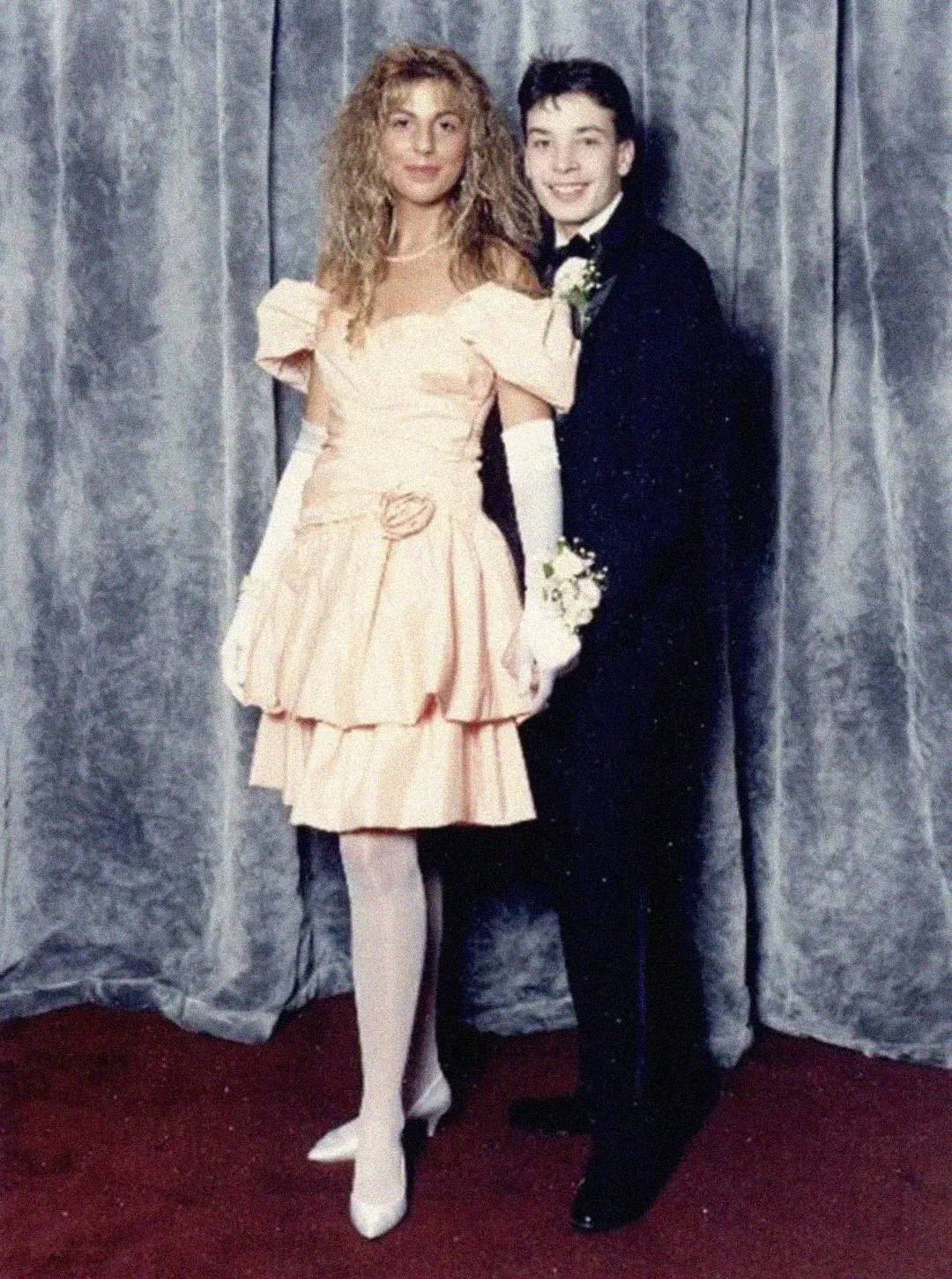 A teenage girl in a pink ruffled dress stands next to a teenage boy in a black tuxedo with a corsage. They pose in front of a gray curtain, smiling, likely at a formal event or prom.