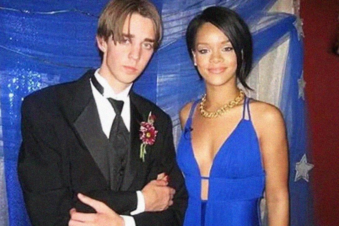 A young man in a black tuxedo stands next to a woman in a deep blue evening gown with a gold necklace. They pose together in front of blue and white drapery decorated with stars.