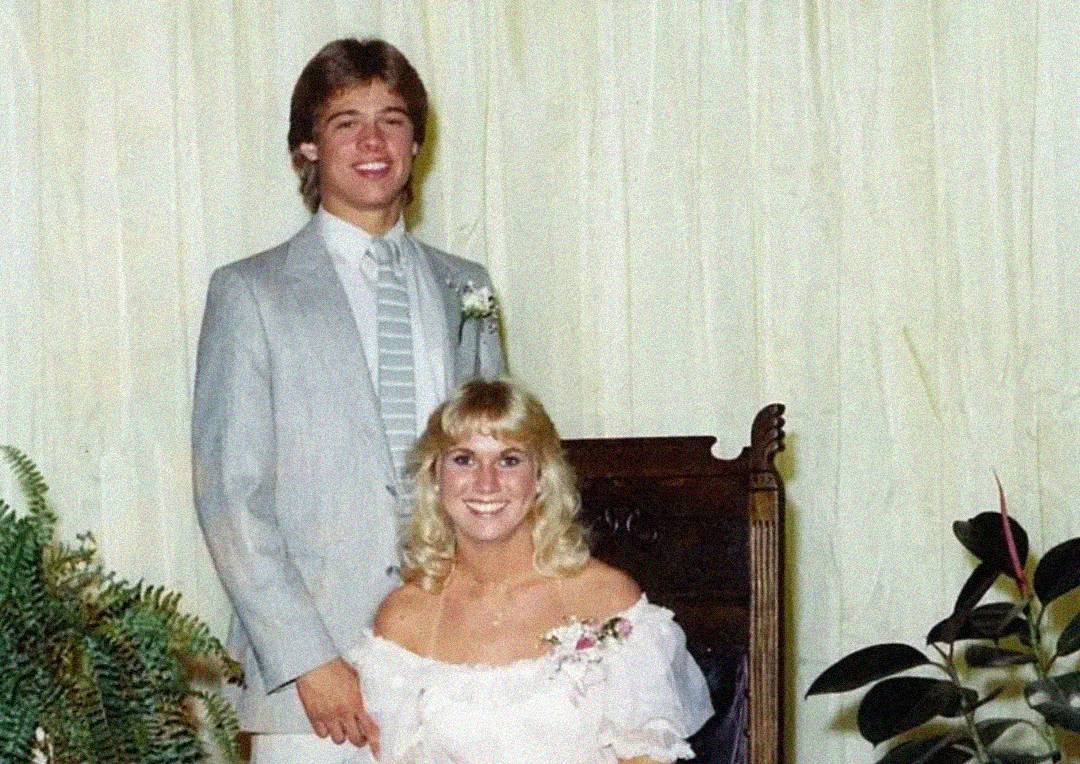 A young man in a light gray suit with a striped tie stands next to a smiling woman in an off-the-shoulder white dress sitting on a dark wooden chair. Both wear corsages, with plants visible in the foreground.