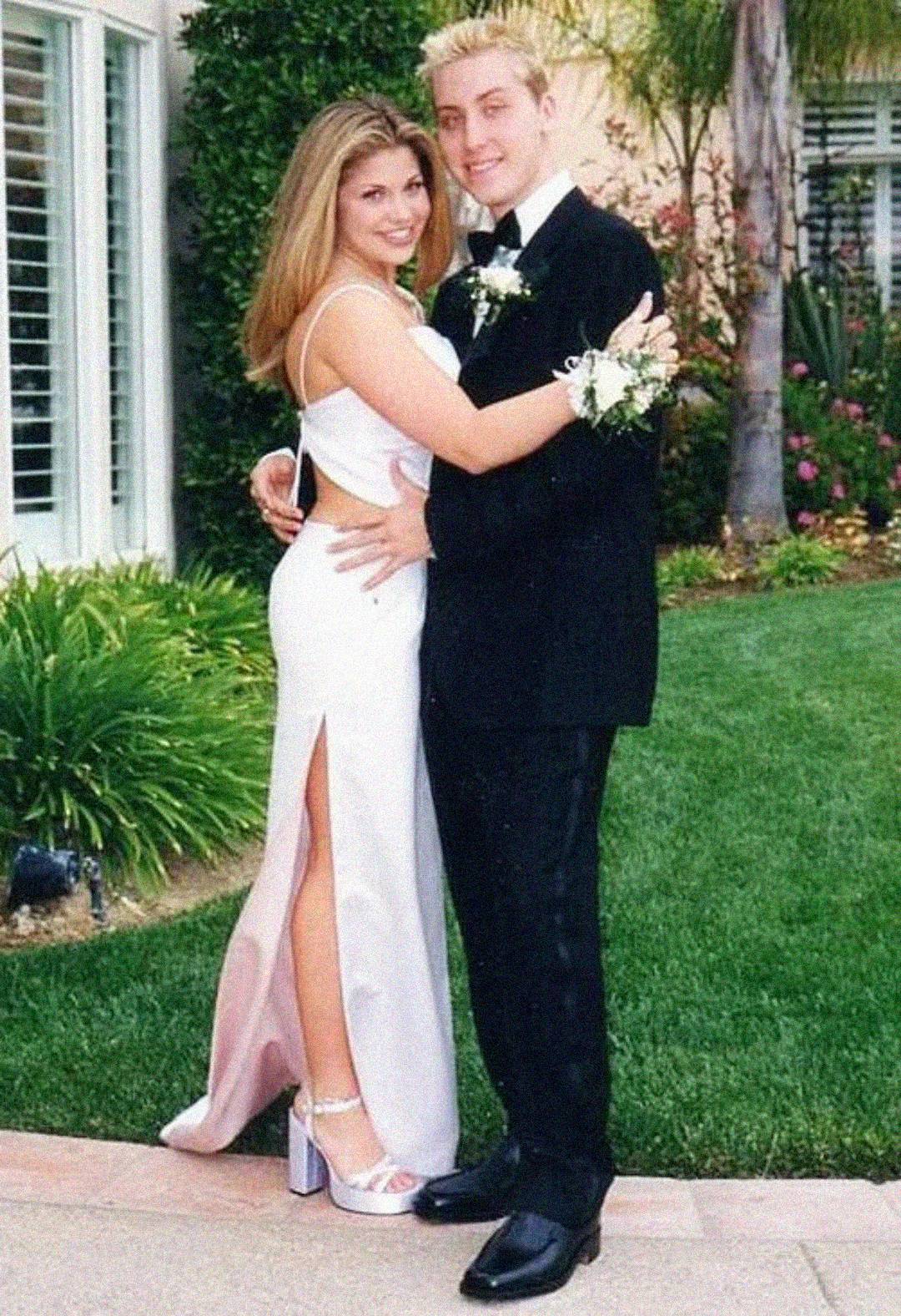 A young couple dressed for prom poses outside; the girl wears a white dress with a high slit and sandals, while the boy wears a black tuxedo. Both have corsages and are smiling, with greenery and a house in the background.