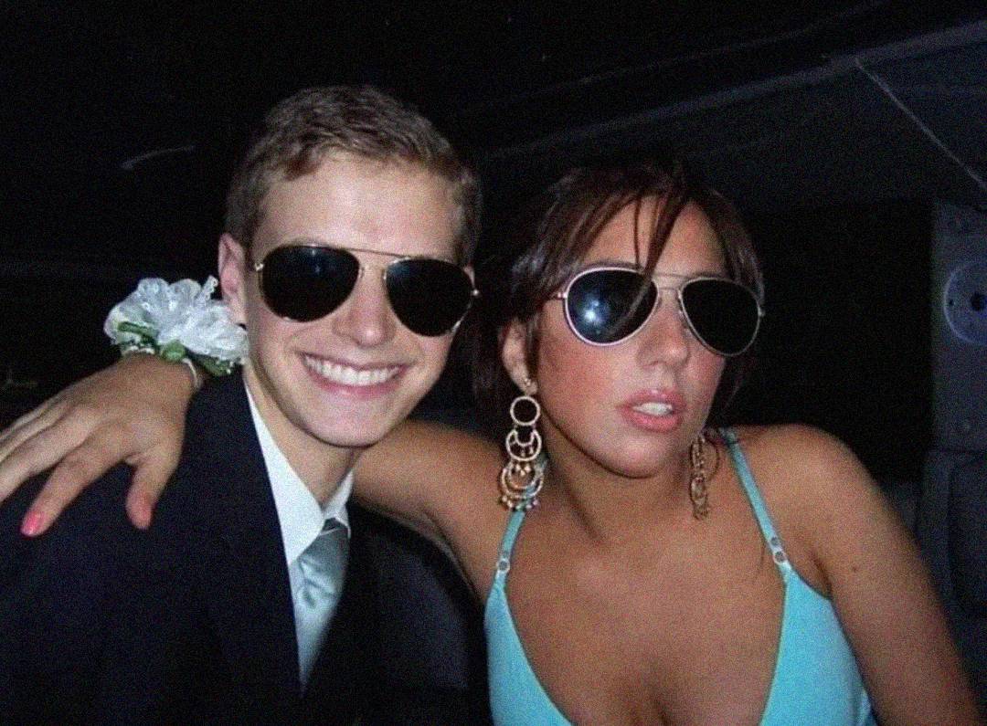 Two young people in formal attire and sunglasses pose together inside a vehicle. The person on the left wears a suit with a boutonniere, while the person on the right wears a blue dress and large earrings. Both are smiling and appear to be at an event.