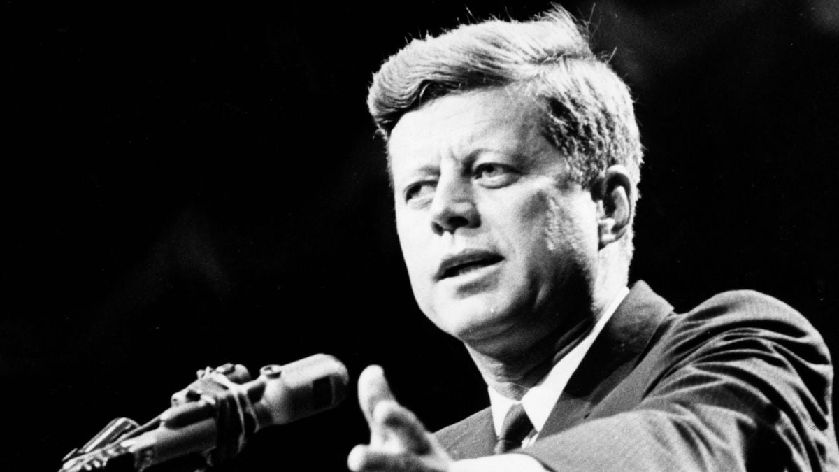A man in a suit speaks into a microphone, gesturing with one hand. The image is in black and white and appears to be from a historical or formal event.