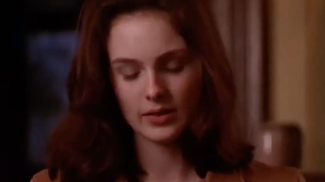A woman with long brown hair looks downward with a neutral expression, wearing a brown jacket indoors. The background is softly blurred.