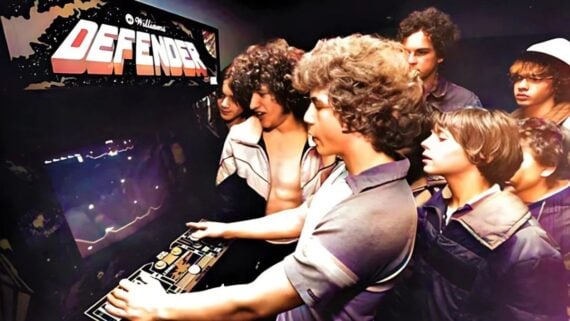 A group of teenage boys intently play and watch the arcade game "Defender" in a dimly lit room, with one boy using the controls while others crowd around, focused on the screen.