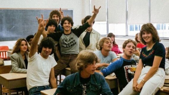 A group of smiling teenagers sits and poses casually in a classroom with desks and a chalkboard, some making hand signs, creating a lively and relaxed atmosphere.