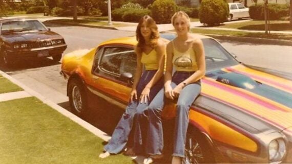 Two women wearing yellow tops and flared jeans sit on the hood of a colorful, retro car parked on a suburban street, with another car parked behind and houses with green lawns in the background.