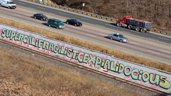 Graffiti spelling &ldquo;Supercalifragilisticexpialidocious&rdquo; in bright colors covers a long concrete barrier beside a highway, with cars and a red dump truck driving on the road above.