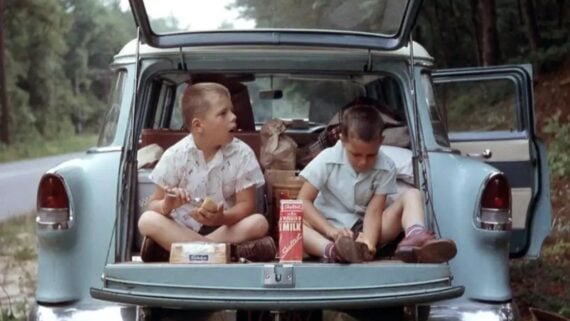 Two young boys sit in the open trunk of a vintage blue car, eating snacks. The car is parked by the roadside with groceries and bags inside. The boys wear light-colored shirts and shorts, surrounded by trees.
