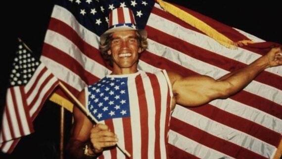 A smiling man in a sleeveless shirt and top hat patterned like the American flag stands in front of a large U.S. flag and holds a smaller flag in one hand, celebrating patriotically.