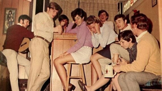 A group of young adults hang out in a cozy, retro room. Some are sitting, laughing, and chatting, while one person plays guitar in the background. The relaxed scene suggests a friendly gathering or house party.