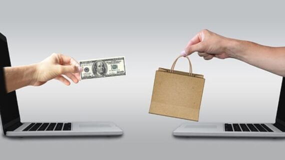 A hand emerges from a laptop screen holding a $100 bill, reaching toward another hand extending from a second laptop holding a small shopping bag, symbolizing online shopping or e-commerce.