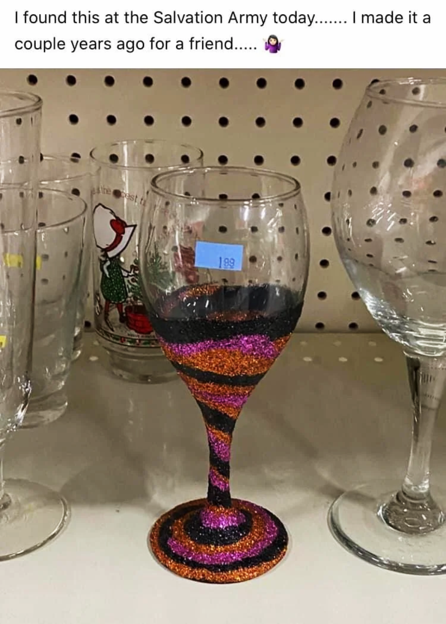 A wine glass with a stem and base decorated in swirling orange, pink, and black glitter is displayed among plain glasses on a thrift store shelf, with a price tag of $1.99 attached.
