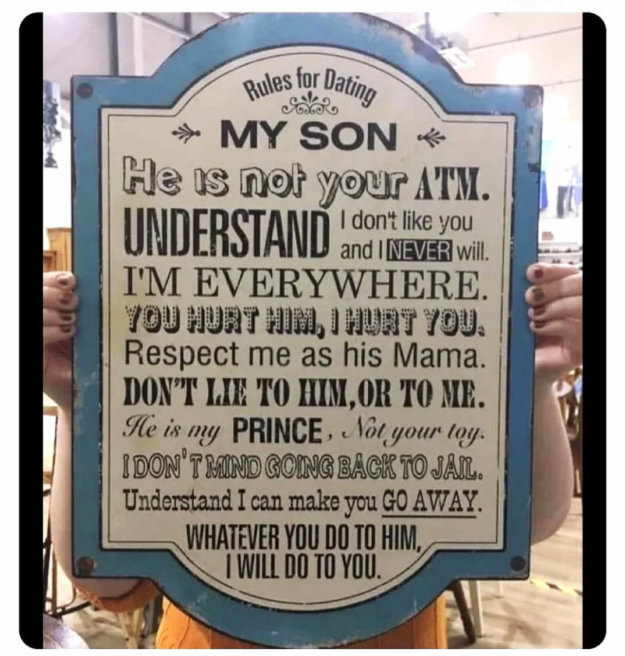 A person holds a decorative sign titled "Rules for Dating My Son," listing protective statements from a mother, warning others not to hurt her son and declaring strong parental boundaries. The sign has a blue border.