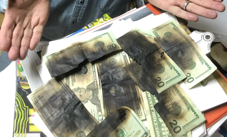 A person with open hands displays several partially burned US twenty-dollar bills spread out on a desk with papers and folders.