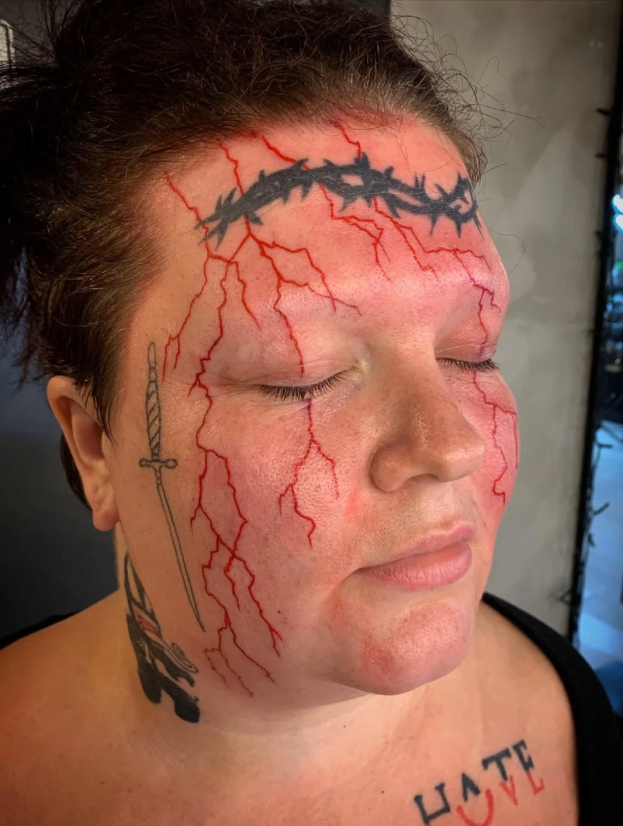 A person with closed eyes has red lightning bolt tattoos across their face, a barbed wire tattoo across their forehead, a sword tattoo on the side of their face, and bold lettering on their chest.