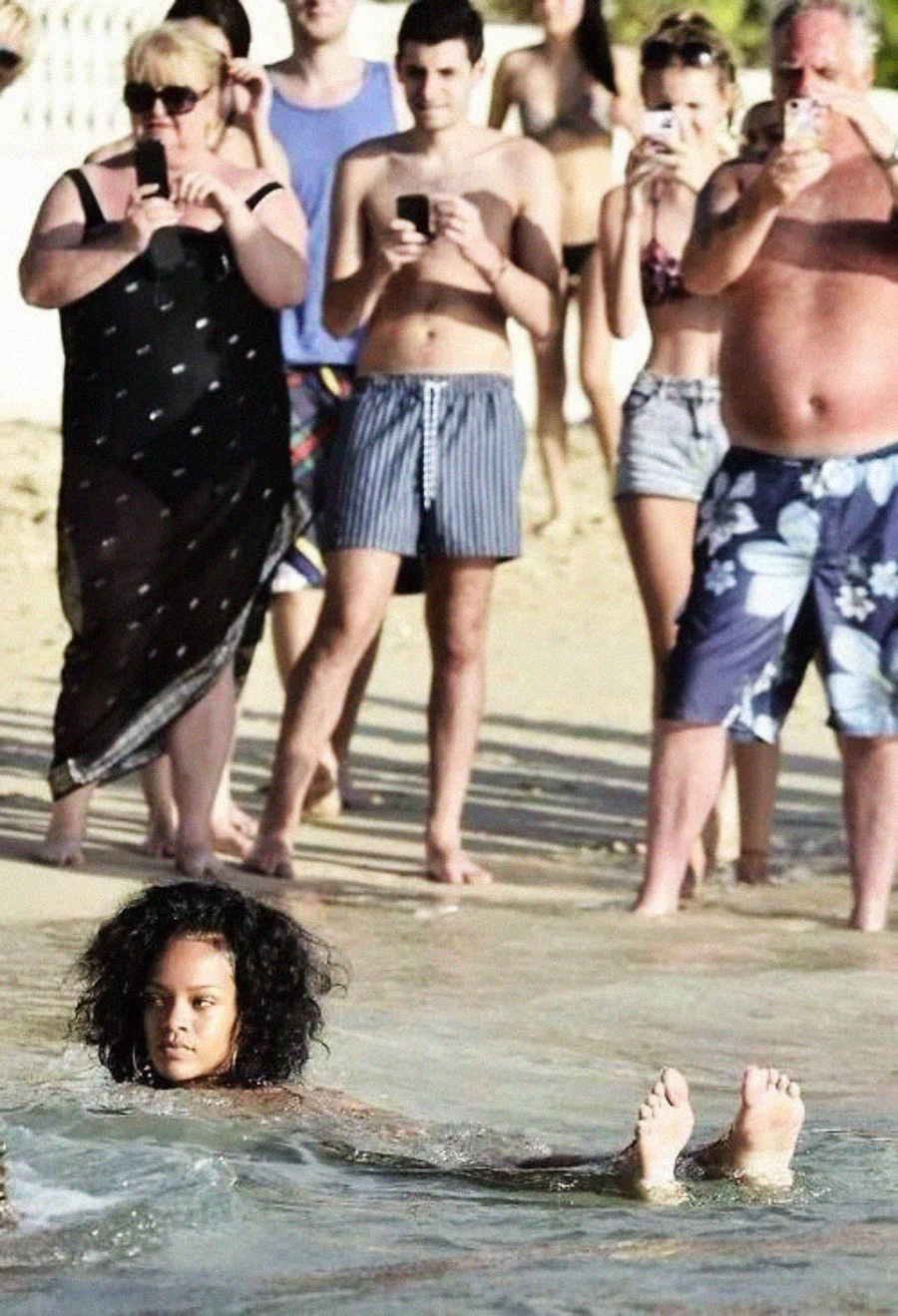 A young woman floats in shallow water at the beach while several people on shore stand watching and taking photos with their phones.