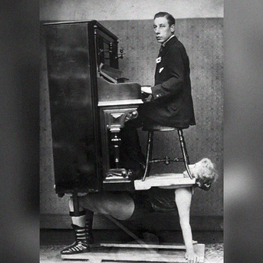 A man in a suit plays an upright piano while sitting on a chair placed on top of the piano. Below him, a woman in stage attire supports the entire piano and chair on her back.