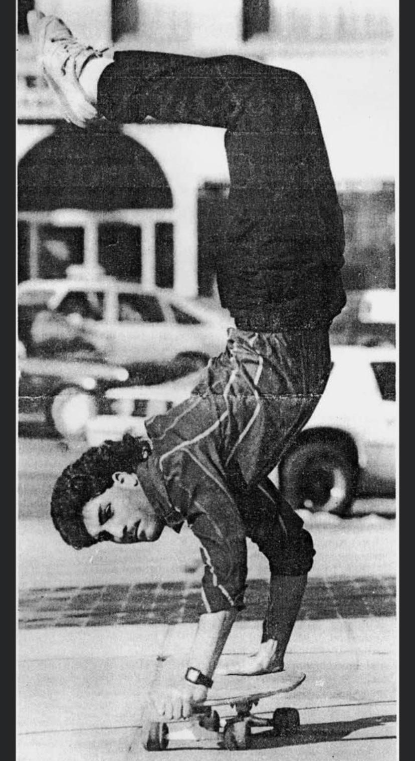 A young man performs a handstand on a skateboard on a city sidewalk, balancing on his hands with his legs straight up in the air. Cars and buildings are visible in the background.