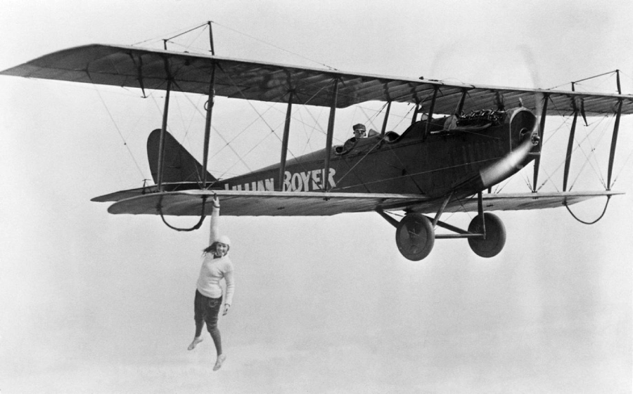 A person hangs by one hand from the wing of a flying vintage biplane with "LYNN ROYER" written on the side, while two pilots sit in the open cockpit above.